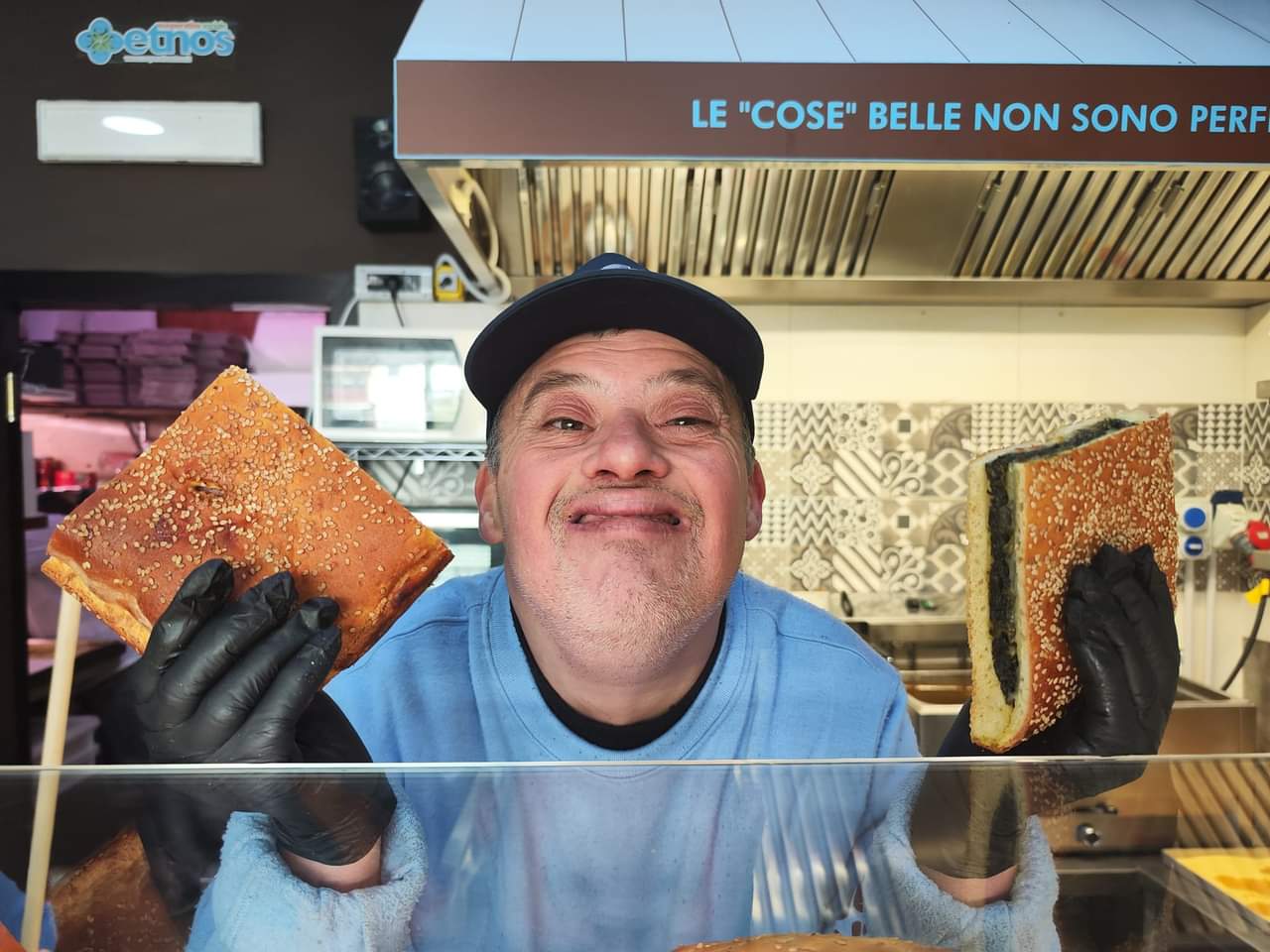 ragazzo sorridente dietro un bancone serve alcuni rustici con verdure; indossa guanti neri in lattice e un cappellino